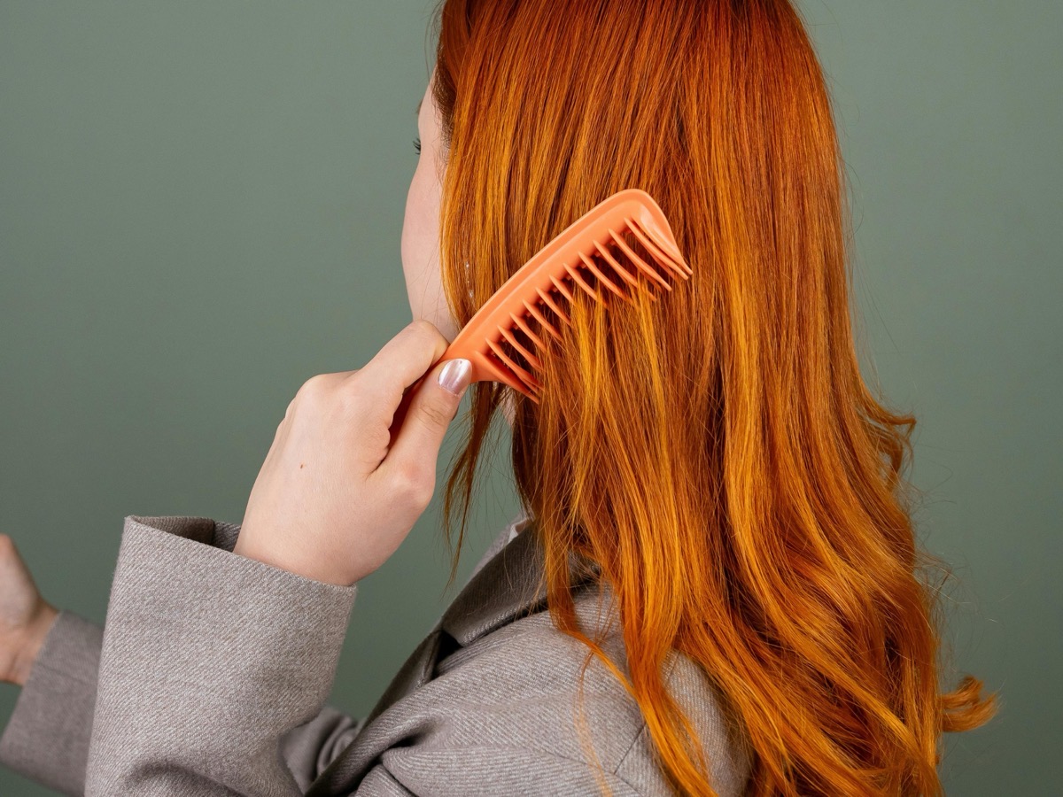 Femme aux longs cheveux roux cuivrés qui les démêle avec un peigne orange à larges dents, sur fond vert sauge